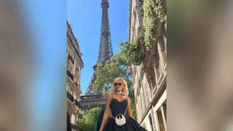 NickyAshcroft poses in a black dress and sunglasses with the Eiffel Tower in the background, standing on a street lined with buildings and trees.