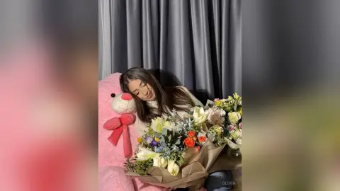 Smiling young woman in a cozy cream sweater holding a large bouquet of colorful spring flowers indoors.