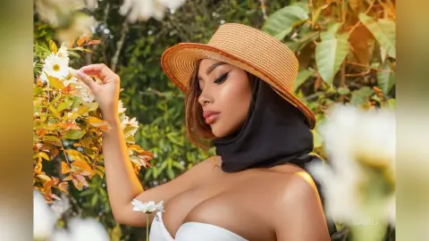 Glamorous woman in a straw sun hat and black scarf posing amidst lush green garden flowers.