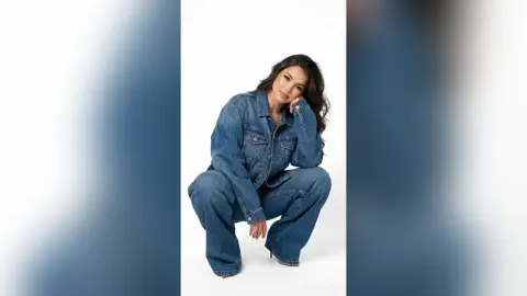 Smiling woman in a blue denim jacket and wide-leg jeans posing against a white background.