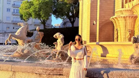 Stylish woman in a white summer outfit posing by a classical stone fountain in warm golden hour sunlight.