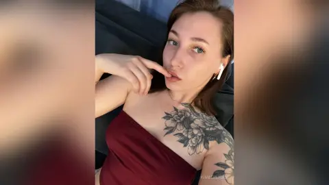 Young woman with floral shoulder tattoo wearing a burgundy strapless top in a relaxed indoor setting.