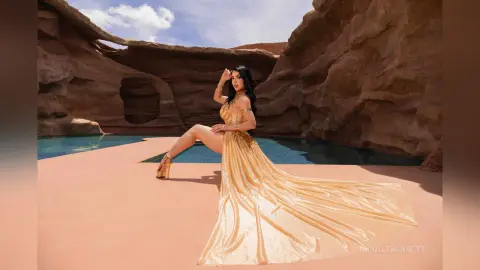 Elegant woman in a flowing gold gown posing on a terrace by a desert rock pool at sunset.