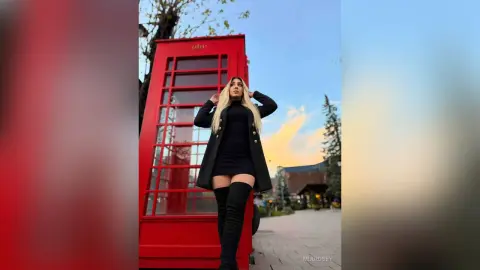 Stylish blonde woman in a black dress and over-the-knee boots posing by a red London telephone booth at sunset.