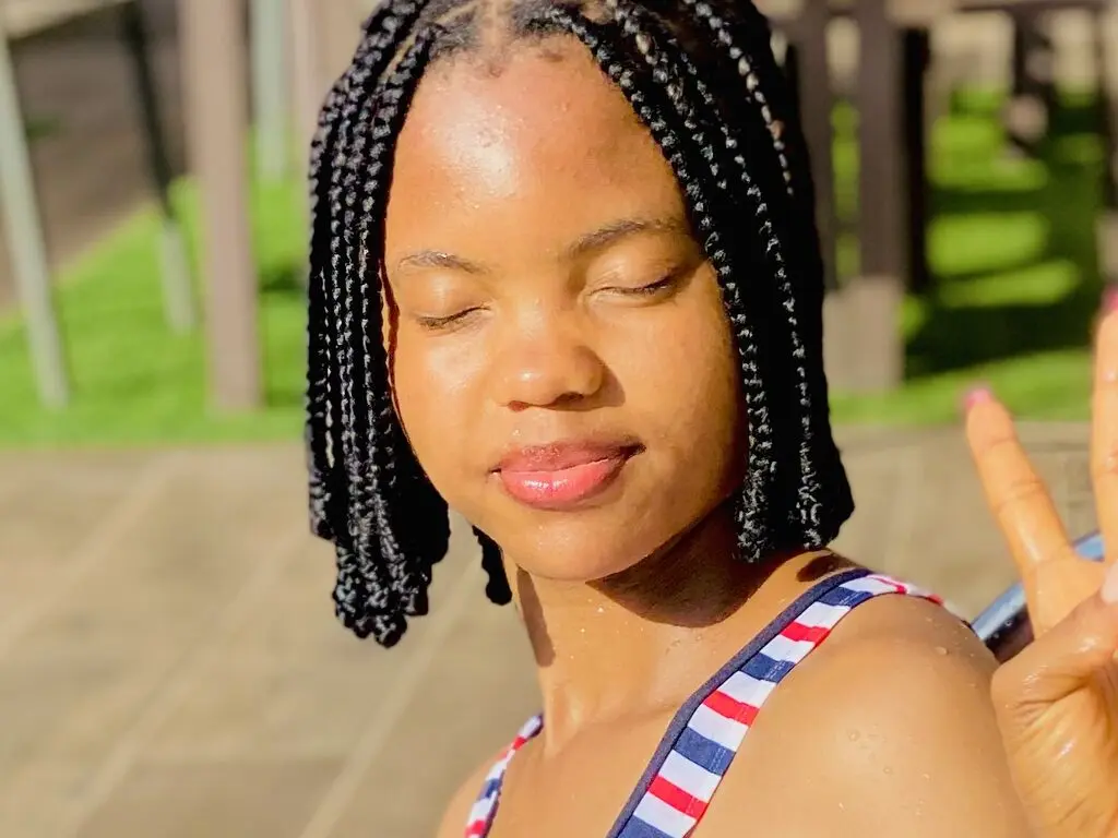 Smiling Black woman with long braids wearing a striped tank top in sunny outdoor setting.