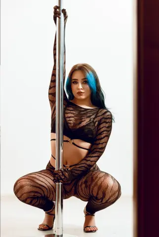 Woman with blue hair in black mesh bodysuit posing confidently against a white studio background.