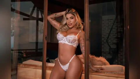 Tattooed woman in white lace lingerie posing confidently in a moody, modern bedroom setting.