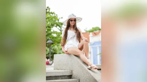 AndreaKalo wearing a white sundress and a wide-brimmed sun hat, sitting on a concrete ledge outdoors with long brown hair and sunglasses.