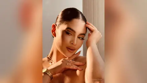 Glamorous woman with slicked-back hair and gold jewelry posing in warm, soft studio lighting.