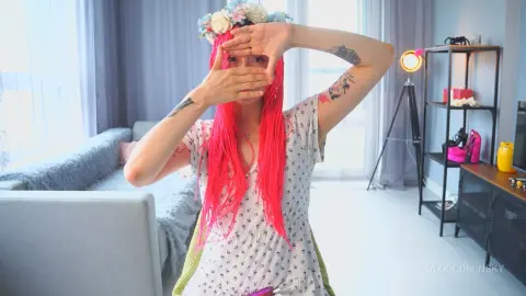 VikkiCominsky frames her face with her hands, showing her bright red hair and a floral headpiece while wearing a white patterned top.