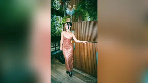 Elegant woman in a copper satin slip dress standing by a wooden wall in a lush, festive indoor setting.