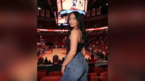 Young woman in a black tank top and blue jeans posing at a crowded indoor basketball arena.