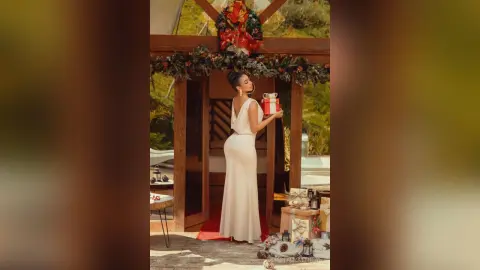 Elegant woman in a white evening gown holding a red gift box in a festive holiday outdoor setting.