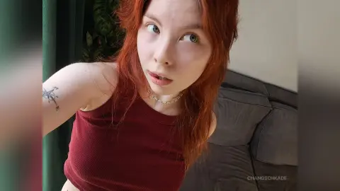 Redheaded woman in a burgundy ribbed crop top posing indoors with a casual, moody expression.