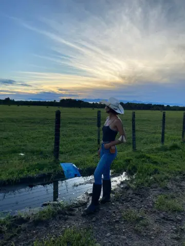CeleGarcia stands in a field by a small pond at sunset, wearing a white cowboy hat, a black tank top, and blue jeans with black boots.
