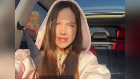Young woman with long brown hair wearing a pink hoodie sitting in a car during golden hour sunlight.