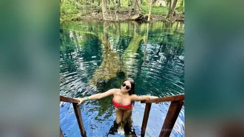 Woman in a red bikini relaxing on a wooden pier by a clear turquoise lake in a lush forest setting.
