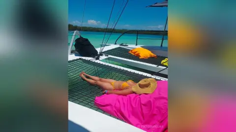 Relaxed woman in a yellow bikini lounging on a pink sunbed on a catamaran deck over turquoise ocean water.