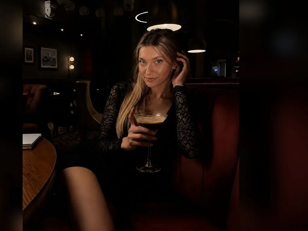 AlmaShannon sits on a red booth in a dark lounge, wearing a black lace dress and holding a cocktail glass.