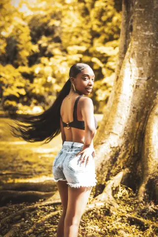 Young woman in denim shorts and black bikini top posing in a sunny, golden forest setting.