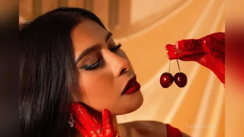 Glamorous woman in red lace gloves holding cherries near her face in warm, moody lighting.
