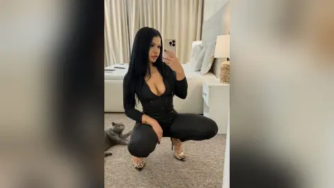 AlmaKane — Woman in a chic black long-sleeve outfit taking a mirror selfie in a modern bedroom with a grey cat.