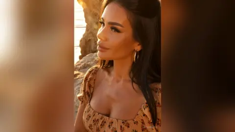 Smiling woman in a floral summer dress enjoying golden hour sunlight by the rocky coast.
