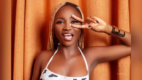 Cheerful Black woman with blonde braids in a white halter top posing playfully against orange curtains.
