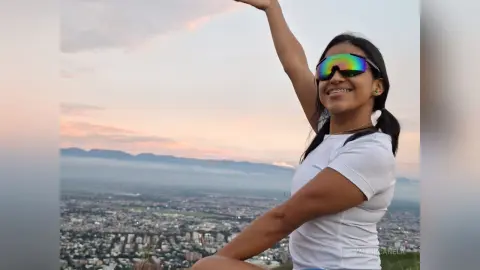 Smiling woman in white t-shirt and sunglasses celebrating at a scenic mountain overlook during sunset.