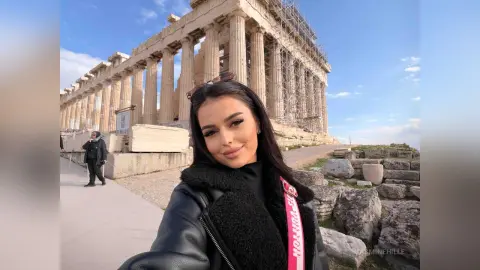 Smiling young woman in a black leather jacket at the ancient Parthenon ruins in Athens under a blue sky.