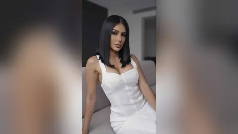 Elegant woman in a white sleeveless dress posing with a confident look in a modern minimalist room.