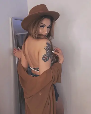 Ameliajade111 — Woman in brown wide-brim hat and cardigan showing shoulder tattoo in a soft indoor setting.