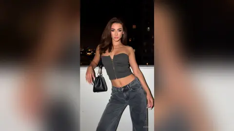 Stylish woman in a dark grey strapless corset top and baggy jeans posing on a city balcony at night.