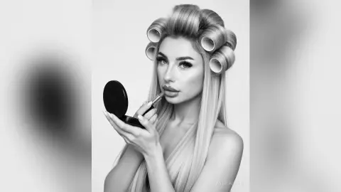Young woman with hair rollers applying makeup in a minimalist studio setting, glamorous beauty portrait.