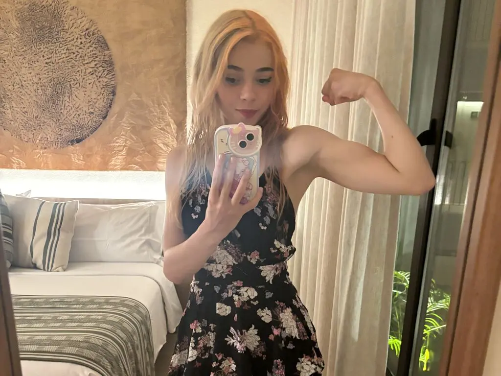 MielVerane poses in a mirror wearing a black floral sundress, flexing her bicep and showing off her blonde hair.