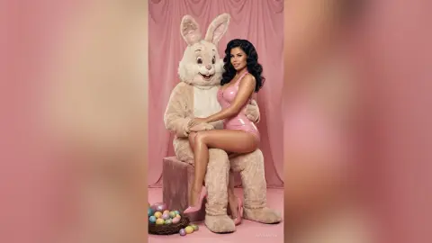 Smiling woman in a pink bodysuit sitting on a large bunny mascot costume in a dreamy pink studio setting.