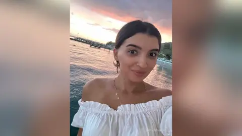 Young woman in a white off-the-shoulder blouse smiling by the ocean at sunset.