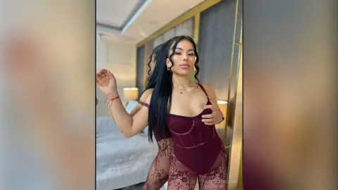 VictoriaCapri poses in a maroon bodysuit and lace tights, showing off her long dark hair and cleavage in a bedroom setting.