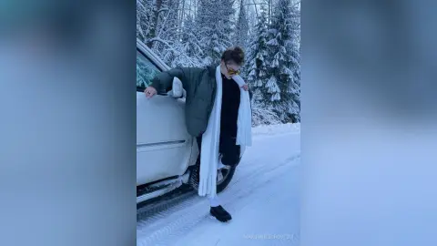Stylish woman in a black outfit and white scarf leaning against a white car in a snowy winter forest setting.
