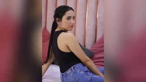 Young woman in a black tank top and blue jeans posing in a cozy pink bedroom setting.