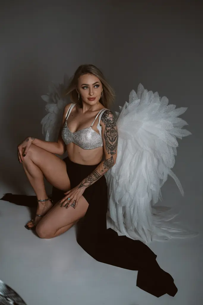Beautiful blonde woman with arm tattoos wearing a white sequin angel costume with large feathered wings.
