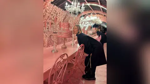 DaisyLin leans over a pink table, sticking her tongue out while wearing a black long-sleeved top in a pink-themed room.
