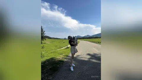 Young woman in a black top and beige skirt holding a dog on a sunny countryside path under a blue sky.