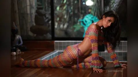 LucyMartin poses on a wooden floor wearing a colorful crochet mesh bodysuit that exposes her breasts and stomach.