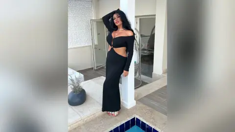 MissyJolie poses outdoors in a long black cutout dress, leaning against a white pillar with one hand in her long dark hair.