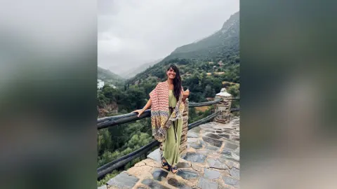 Smiling woman in a green maxi dress and patterned shawl standing on a stone balcony overlooking a misty mountain valley.