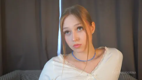 Melancholic young woman with blonde hair wearing a white lace blouse and blue beaded necklace indoors.