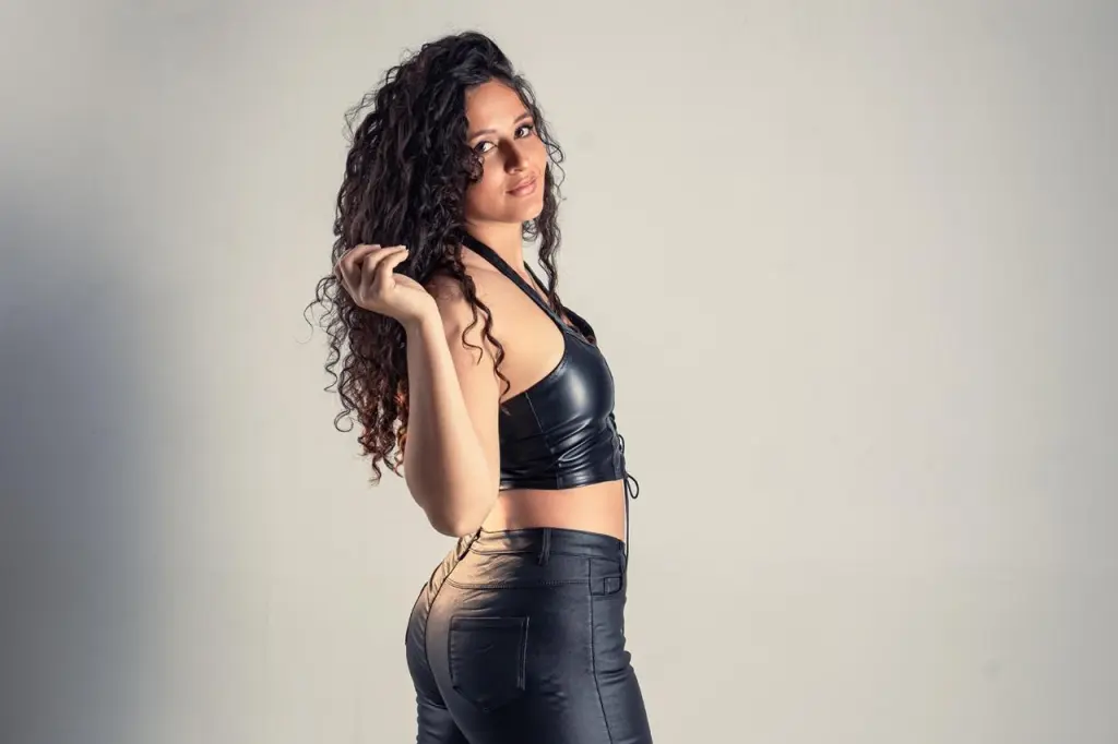 Dannaloves poses in profile, wearing a black leather bra and matching bottoms, showing her long curly dark hair against a plain light background.