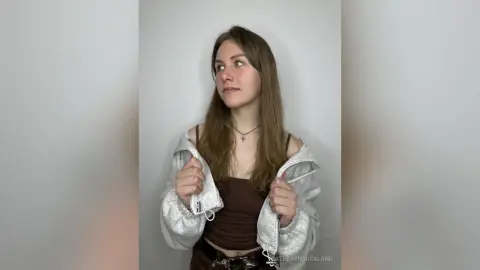 CatherynBuckland wears a white zip-up hoodie over a brown tank top, looking off to the side against a plain white background.