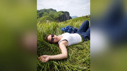 MilaSwan — MilaSwan lies in tall green grass on a mountain hillside wearing a white tank top and sunglasses.
